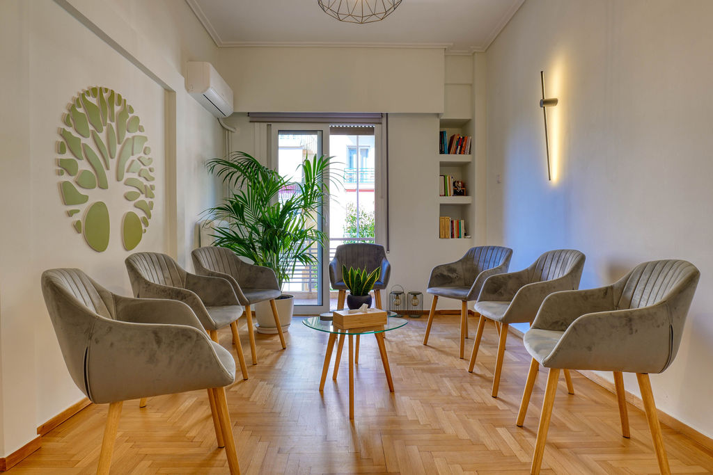 Χώρος αναμονής με ξύλινο πάτωμα, γκρι πολυθρόνες, φυτά εσωτερικού χώρου και διακόσμηση τοίχου με σχέδιο δέντρου.