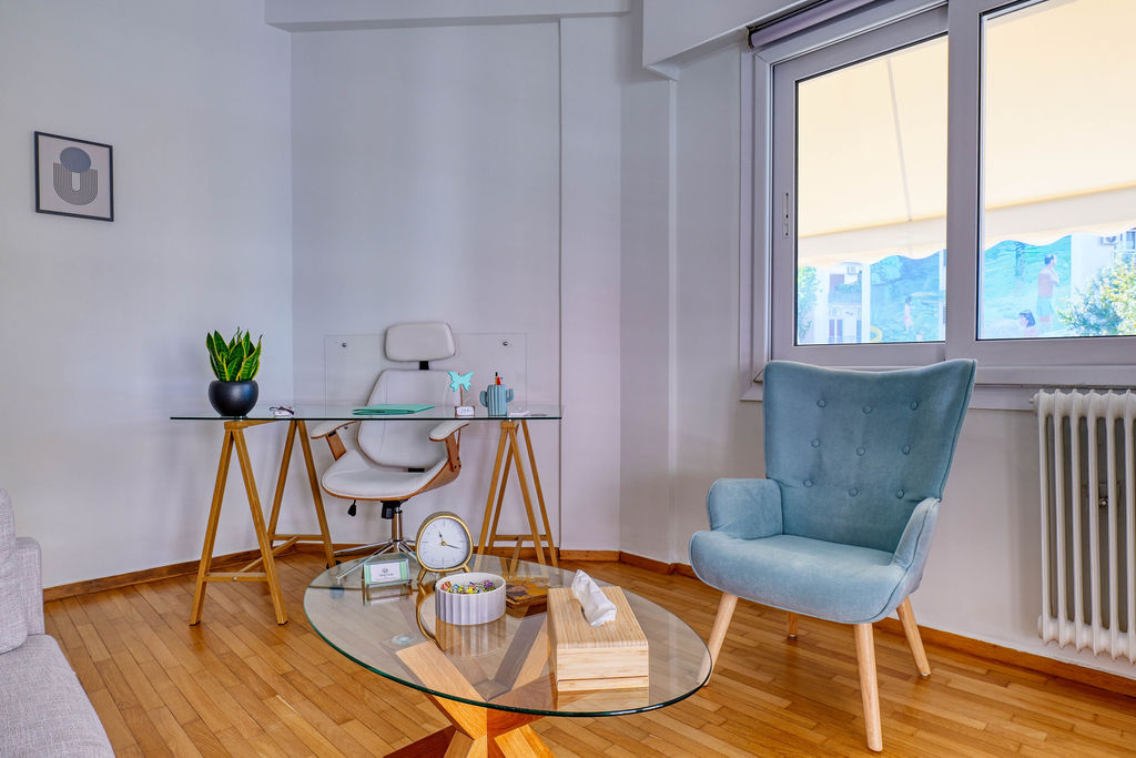 Εσωτερικός χώρος με ξύλινο πάτωμα, γραφείο, γυάλινο τραπεζάκι και γκρι πολυθρόνα.