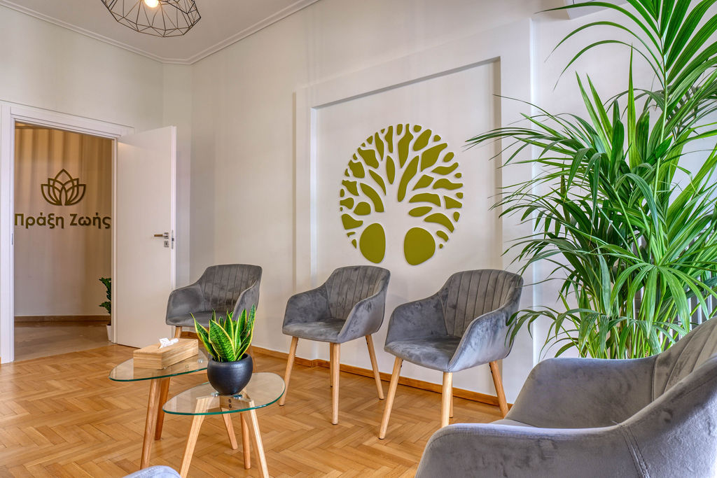 Φωτεινός χώρος αναμονής με ξύλινο πάτωμα, γκρι πολυθρόνες, φυτά εσωτερικού χώρου και διακόσμηση τοίχου με σχέδιο δέντρου. Στο βάθος διακρίνεται λογότυπο με ελληνική επιγραφή "Πράξη Ζωής".
