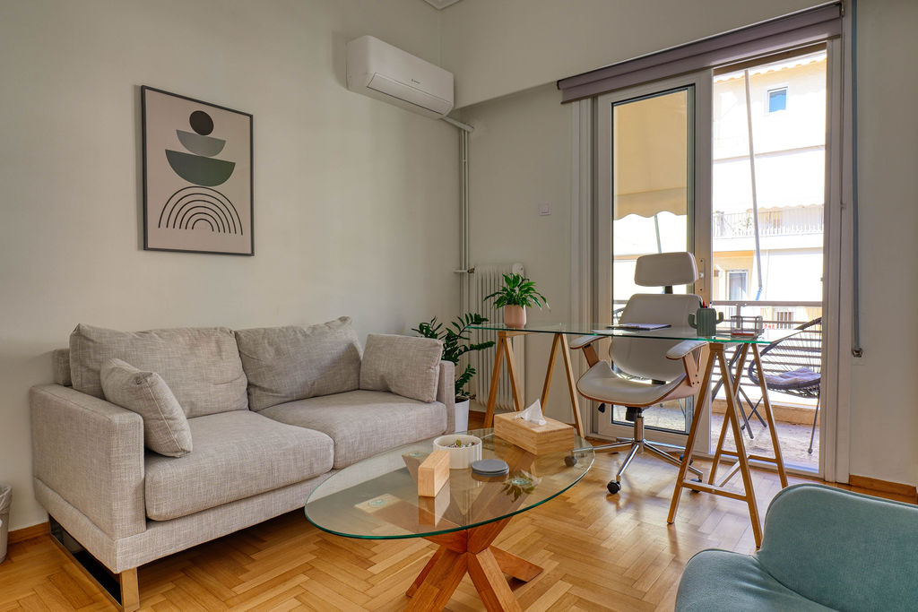 Εσωτερικός χώρος με ξύλινο πάτωμα, γκρι καναπέ, γυάλινο τραπεζάκι και γραφείο.